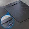 A close-up view of a black, slate-look shower tray with blue arrows indicating water flow towards the drain. A magnified inset shows water and debris near the drain. Produktbild-Vorschau