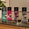 Four boxes of Tchibo Cafissimo coffee capsules are displayed on a dark surface, with a shot of espresso in a glass in front of them. Coffee beans are scattered on the surface. Produktbild-Vorschau