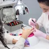 A nail technician applies nail polish to a client's fingernails under a UV lamp. A case filled with nail polish bottles and nail tools is visible. Produktbild-Vorschau