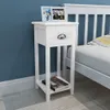 A white bedside table with a drawer and a shelf, placed next to a bed with a white headboard and a striped pillow. The wall behind is blue. Podgląd zdjęcia produktu