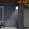 A modern gray front door with a sleek handle, illuminated by a white LED wall light. A potted lavender plant sits to the left. Produktbild-Vorschau