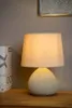 A table lamp with a beige fabric shade and a textured beige base sits on a wooden surface. A decorative box is to the left of the lamp. A framed artwork hangs on the wall behind. Náhled obrázku produktu