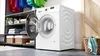 A white Bosch washing machine with its door open, placed in a laundry room with shelves of folded towels and baskets. Produktbild-Vorschau