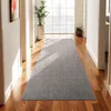 A long, gray runner rug is placed on a polished wooden floor in a hallway. Sunlight streams in from a large window at the end of the hallway, casting shadows on the rug. There are two potted plants in the hallway, one large and one small, and a framed piec Produktbild-Vorschau