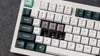 A close-up view of a white mechanical keyboard with dark green and white keycaps. Some keycaps are removed, revealing purple switches. Produktbild-Vorschau