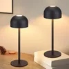 Two black cordless LED table lamps are displayed on a wooden surface, one in the foreground and one slightly behind and to the right. A pinecone and a wooden coaster are visible to the left of the foreground lamp. A stack of books is placed under the lamp  Náhľad obrázku produktu