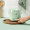 A light green mini rice cooker is on a wooden cutting board, with a hand pressing a button on the rice cooker. Produktbild-Vorschau