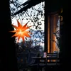 An orange star lantern hangs from a tree branch, with a table and chairs set up on a balcony in the background, illuminated by a candle. Produktbild-Vorschau