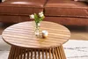 A round wooden coffee table with a vase of flowers and a cup on it, in front of a brown leather sofa. Produktbild-Vorschau