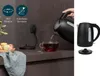 A person is pouring hot water from a black electric kettle into a glass cup of tea. The scene is set on a kitchen counter with macarons in a bowl. Produktbild-Vorschau