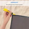 A hand is holding a yellow stake to anchor a gray beach blanket to the sand. Produktbild-Vorschau
