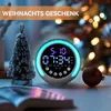 A digital alarm clock with RGB night light is displayed on a table next to a small decorative Christmas tree and a Christmas ornament, with a blurred background of Christmas lights. Produktbild-Vorschau