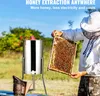 A beekeeper in protective gear holds a honeycomb frame next to a manual honey extractor in an outdoor setting. Podgląd zdjęcia produktu
