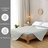 A bed with a light gray quilted blanket draped over it, with two white pillows at the head of the bed. The scene is set in a bedroom with curtains and a clock on the wall. Náhled obrázku produktu