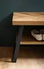 A close-up of a wooden shoe bench with a herringbone pattern on the top, a black metal leg, and a shelf with shoes on it. The background is a dark green wall and a wooden floor. Produktbild-Vorschau