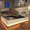 A white upholstered bed with its storage compartment open, revealing folded linens and pillows. The bed has LED lighting around its base and a tufted headboard. The room is dimly lit, with a window showing a night sky. Náhled obrázku produktu