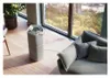 An air purifier is placed on a wooden floor next to a large window with a city view, and a sofa with pillows. Podgląd zdjęcia produktu