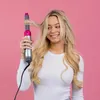 A young woman with blonde hair is holding a hair styling tool against a pink background. Produktbild-Vorschau