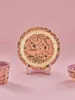 A plate with a jungle print is displayed on a pink background, with a cup and bowl partially visible on the sides. Produktbild-Vorschau