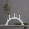 A white arched candle holder with seven lit electric candles sits on a wooden table, with a sprig of greenery hanging from above and mossy decorations on the table. Produktbild-Vorschau