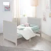 A baby bed with bedding and pillows in a room with artwork on the wall and a window with sheer curtains. Produktbild-Vorschau