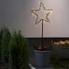 A star-shaped LED light decoration is placed in a pot with apples and greenery, next to some evergreen bushes and a basket of firewood. Podgląd zdjęcia produktu