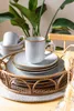 A set of stoneware dishes and mugs are arranged on a rattan tray, with a lush green plant in the background. Produktbild-Vorschau