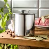 A stainless steel cooking pot with a lid, placed on a wooden surface, with ingredients like garlic, peppercorns, bay leaves, and raw meat in the background. Podgląd zdjęcia produktu