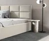 A close-up view of a bed with a beige upholstered headboard and a bedside table with a lamp. Produktbild-Vorschau