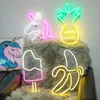A collection of neon lights shaped like a unicorn, pineapple, popsicle, and banana, arranged on a furry rug and a sequined pillow. Náhled obrázku produktu