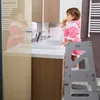 A toddler is brushing her teeth in a bathroom, standing on a grey step stool. Produktbild-Vorschau