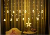 A curtain of fairy lights with round bulbs hangs in front of a yellow curtain. A star-shaped light and a small radio are on a table below. Náhled obrázku produktu