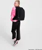 A young girl wearing a pink tie-dye sweatshirt and black pants stands facing away from the camera, wearing a black backpack. Produktbild-Vorschau
