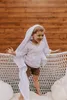 A young boy is standing on a white woven bench, wrapped in a white cloth, with a wooden fence in the background. Produktbild-Vorschau