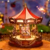 A miniature carousel with horses, illuminated by LED lights, set against a blurred background of lights and greenery. Produktbild-Vorschau