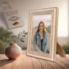 A framed photo of a woman is displayed on a wooden table next to a vase with greenery. A tag with German text and the German flag is also visible. Produktbild-Vorschau