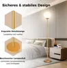 A modern floor lamp with a gold-colored metal pole and a white globe shade is shown in a bedroom setting next to a nightstand and bed. Podgląd zdjęcia produktu