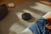 A black robot vacuum cleaner is cleaning a rug with scattered debris. A couch and a cushion are visible in the foreground. Produktbild-Vorschau