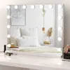 A vanity mirror with lights is placed on a white surface in front of a bed with white pillows and a white blanket. Produktbild-Vorschau