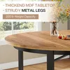 Close-up of a round wooden dining table with black metal legs, set near a window with greenery outside. There are two apples and a vase with flowers on the table. Podgląd zdjęcia produktu