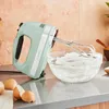 A mint green KitchenAid hand mixer is shown in use, whipping cream in a glass bowl on a kitchen counter. Eggs and eggshells are visible in the foreground. Podgląd zdjęcia produktu