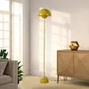 A yellow floor lamp is placed in a living room next to a sofa and a wooden cabinet. Produktbild-Vorschau