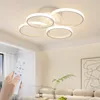 A hand holding a remote control is in the foreground, with a modern, multi-ringed ceiling light fixture illuminated above. The background shows a living room setting with a sofa and artwork. Podgląd zdjęcia produktu