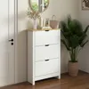 A white shoe cabinet with a wooden top, placed next to a white door and a large potted plant. Produktbild-Vorschau
