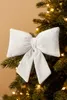 A white velvet bow ornament is attached to a Christmas tree branch, with fairy lights visible in the background. Produktbild-Vorschau