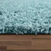A close-up view of a fluffy, light blue shaggy rug placed on a wooden floor. Produktbild-Vorschau