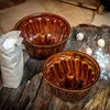 Two brown ceramic bundt cake molds are displayed on a rustic wooden surface, with a bag of white powder (likely flour) and some dried seed pods nearby. Voir les images du produit