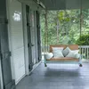 A porch swing with cushions is shown on a wooden porch, with trees and foliage in the background. Náhľad obrázku produktu