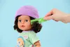 A doll wearing a purple hat and green shirt, being combed with a green comb. Produktbild-Vorschau