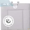 A light gray baby changing mat with a teddy bear face design at the top. A circular certification badge is on the left side. Náhľad obrázku produktu